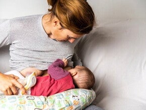 Kobieta siedząca na kanapie karmi niemowlę piersią, jednocześnie trzymając w dłoni butelkę z mlekiem, co ilustruje karmienie mieszane.