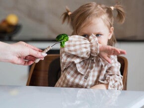 Dziecko odmawiające jedzenia – mała dziewczynka odsuwa ręką brokuł podawany na widelcu, obraz ilustrujący trudności w karmieniu u dzieci (PFD/ARFID)