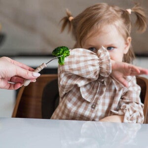 Dziecko odmawiające jedzenia – mała dziewczynka odsuwa ręką brokuł podawany na widelcu, obraz ilustrujący trudności w karmieniu u dzieci (PFD/ARFID)