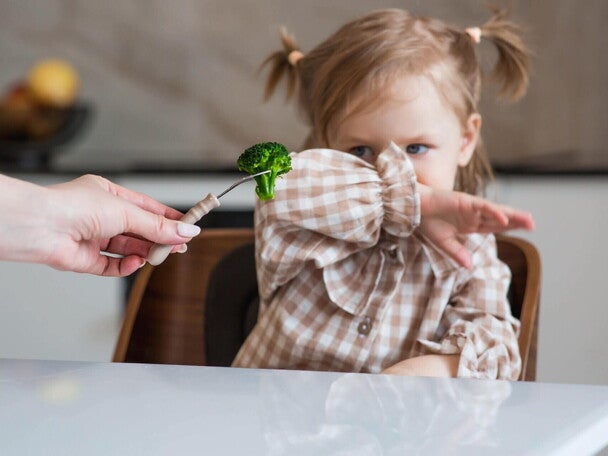 Dziecko odmawiające jedzenia – mała dziewczynka odsuwa ręką brokuł podawany na widelcu, obraz ilustrujący trudności w karmieniu u dzieci (PFD/ARFID)